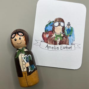 Puede incluir: Una muñeca de madera pintada para parecerse a Amelia Earhart, con una chaqueta de cuero marrón, una bufanda verde y un casco de aviador de cuero marrón. La muñeca sostiene un mapa. Una tarjeta con un dibujo de Amelia Earhart y el texto "Amelia Earhart" está en el fondo.