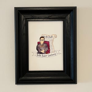 May include: A framed illustration of Ruth Bader Ginsburg, a Supreme Court Justice, sitting in a chair with a scroll of the US Constitution behind her. The scroll reads "We the People". The illustration is in a black frame.