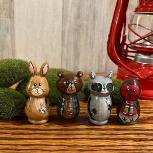 May include: Four hand-painted wooden animal figurines: a rabbit, a bear, a raccoon, and an owl. The rabbit is tan with pink ears, the bear is brown, the raccoon is grey, and the owl is red. Each figurine is approximately 7.6 cm tall.