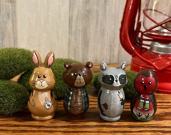 Puede incluir: Cuatro figuras de animales de madera pintadas a mano: un conejo, un oso, un mapache y un búho. El conejo es marrón claro con orejas rosas, el oso es marrón, el mapache es gris y el búho es rojo. Cada figura mide unos 7,6 cm de altura.