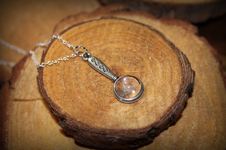 May include: A silver necklace with a magnifying glass pendant. The pendant has a decorative design on the handle and a clear glass lens.
