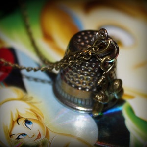 May include: A vintage-style thimble pendant necklace. The thimble is silver-toned with a textured surface and a gold-colored chain. Small gold-colored acorn charms dangle from the thimble. The background features a colorful illustration of a cartoon character.