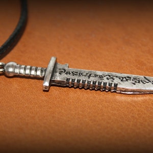 May include: A silver-toned pendant necklace featuring a detailed dagger design with an inscription on the blade. The necklace is on a black leather cord.