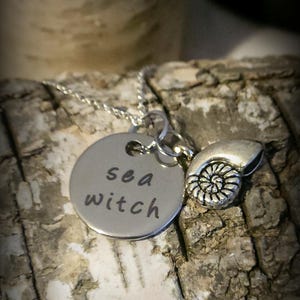 May include: A silver-toned necklace featuring a round metal pendant stamped with the text sea witch in lowercase letters, accompanied by a small, detailed silver-toned charm shaped like a spiral ammonite fossil shell on a thin metal chain.