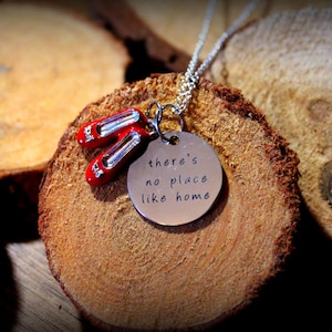 May include: A silver-toned necklace featuring a round metal pendant engraved with the text 'there's no place like home' and a pair of miniature red shoes attached to the jump ring, suspended from a delicate silver-toned metal chain.