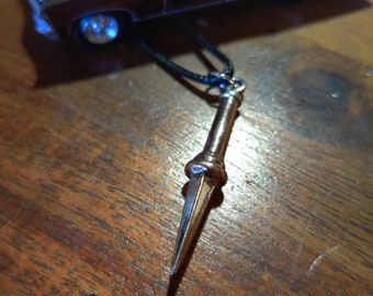 May include: A silver-colored pendant shaped like a spear point hangs from a black cord. The pendant is about 4 inches long and has a pointed tip. The pendant is lying on a wooden surface.