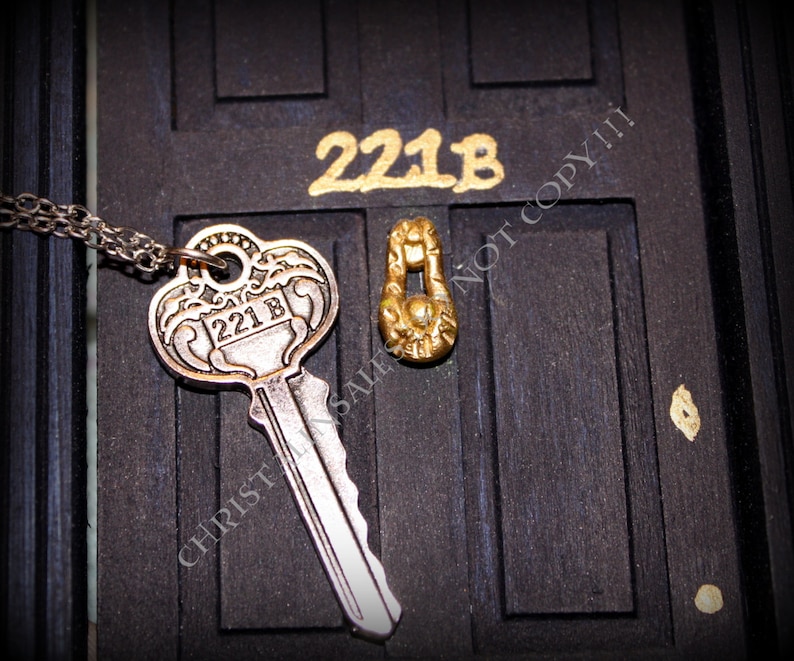 May include: A silver key with the number "221B" engraved on it hangs from a silver chain. The key is in front of a black door with a gold door knocker and the number "221B" written in gold paint.