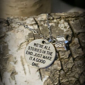 May include: A silver-toned metal necklace featuring a round pendant with the engraved text WE'RE ALL STORIES IN THE END. JUST MAKE IT A GOOD ONE. and a small charm shaped like an open book, suspended from a delicate silver-toned metal chain.