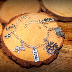 May include: Silver charm bracelet with various charms, including a typewriter, a sun with a pentagram, a goat, and a tag that reads "J & S". The bracelet is displayed on a wooden slice.