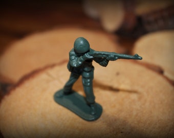 May include: A green plastic toy soldier holding a rifle, standing on a wooden surface.