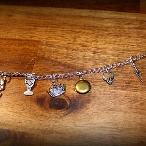 May include: A silver charm bracelet with various charms, including a snake, a goblet, a crown, a locket, a ring, a lightning bolt, and a book. The bracelet is on a wooden surface.