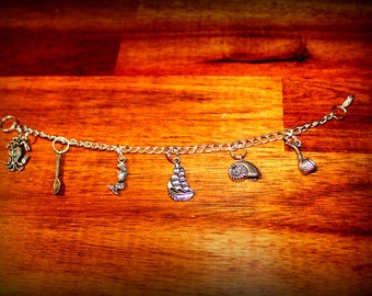 May include: A silver charm bracelet with various charms, including a ship, a mermaid, a seashell, a spoon, and a pipe. The bracelet is on a wooden surface.