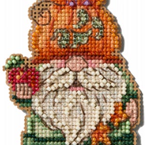 May include: A cross-stitch decoration in the shape of a gnome, featuring a pumpkin hat and beard. The gnome's hat is orange with green leaves, and the beard is white. The gnome is holding a bouquet of flowers. The overall color scheme is orange, green, and white.