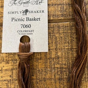 May include: A skein of brown embroidery floss with a tag that reads "The Gentle Art SIMPLY SHAKER Picnic Basket 7060 COLORFAST." The floss is a rich, earthy brown color, and the tag is cream-colored with a circular hole at the top.