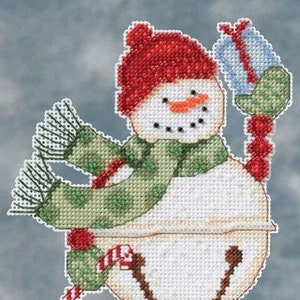 May include: Cross-stitch snowman ornament wearing a red hat and green scarf, holding a blue gift. The snowman has a carrot nose and a smiling face. A large bell is at the bottom, with a red beaded strand. Festive holiday decor.