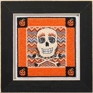 May include: A cross-stitch pattern featuring a cartoon skull with a toothy grin, wearing a pumpkin hat and surrounded by orange and grey chevron patterns. The skull is holding crossed bones.