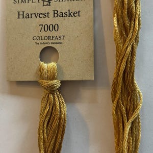 May include: A skein of gold-colored embroidery floss from The Gentle Art, labeled "Harvest Basket 7000" and "Colorfast." The floss is attached to a tan card with a hole for hanging. The floss is a twisted bundle.