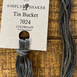 May include: A skein of gray and black embroidery floss labeled "Tin Bucket 7024" by The Gentle Art. The colorfast thread is tied with a knot and attached to a cream-colored tag with a hole for hanging. The background is a wooden surface.