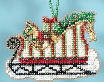 May include: A beaded Christmas ornament depicting a red sleigh filled with presents, a teddy bear, and a horse. The sleigh is decorated with green and white stripes and has a green garland on top.