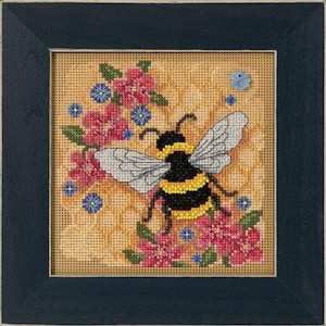 May include: Cross-stitch artwork of a bumblebee with yellow and black stripes, surrounded by pink flowers and blue accents. The bee has gray wings and is set against a honeycomb background. The artwork is framed in a dark blue wooden frame.