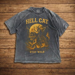 May include: Dark grey t-shirt with a distressed print featuring the text "HELL CAT" above an illustration of a skeleton riding a tiger in front of a full moon. The text "STAY WILD" is below the illustration.
