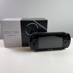 以下が含まれることがあります： ピアノブラック仕上げの黒いソニー製携帯ゲーム機、PlayStation Portable。オリジナルの箱の前に配置されています。箱にはPSPのロゴ、PlayStation Portableの文字、およびモデル名Piano Black PSP-3000が記載されています。