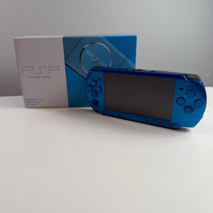 Pode incluir: Uma consola de jogos portátil azul vibrante com ecrã preto, painel direcional e botões de ação. A consola está posicionada em frente à sua caixa de embalagem original branca e azul, que apresenta o texto PSP e PlayStation Portable.