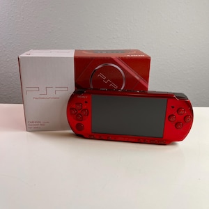 May include: A radiant red Sony PlayStation Portable handheld gaming console with a black screen and control buttons, positioned in front of its original white and red retail box labeled with the text PSP PlayStation Portable Carnival Colors.