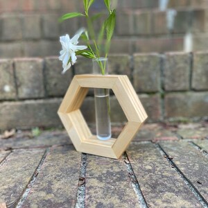May include: A wooden hexagonal vase with a glass test tube in the center. The vase is made of light wood and has a natural finish. A single white flower is in the test tube.