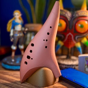 May include: A pink ocarina with multiple finger holes, resting on a brown stand. The ocarina is a musical instrument, and the image also features a figurine of a character and a mask from the Legend of Zelda video game series.