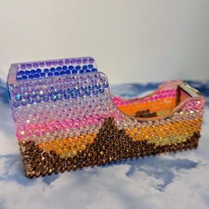 May include: A tape dispenser covered in rows of small, sparkling rhinestones arranged in a gradient pattern. The colors transition from dark blue at the top to purple, pink, orange, yellow, and brown at the base, creating a vibrant, multi-colored effect.