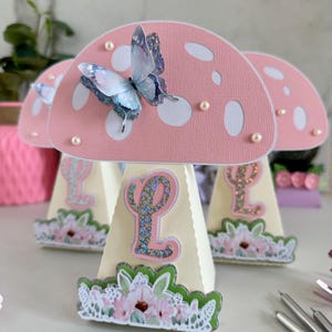 Puede incluir: Tres cajas de regalo de fiesta en forma de hongo rosa y blanco con un diseño de mariposa. Cada caja tiene una base cónica blanca con un diseño floral y una letra L brillante.