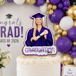Puede incluir: Un adorno para pastel de graduación con una mujer con birrete y toga morados sosteniendo un diploma. El adorno tiene la palabra "Graduation" y el año "2026". El pastel es blanco con glaseado decorativo.