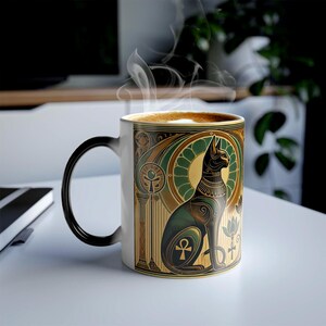 May include: A white ceramic mug with a black handle, showcasing an Egyptian cat design in gold, green, and black. The mug contains a hot drink, with steam rising. The design incorporates an ankh symbol and lotus flowers. The mug is likely 10-12 cm tall.