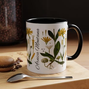 May include: White ceramic mug with a black rim and handle. The mug features a botanical illustration of Arnica Montana flowers in yellow and green on a white background.