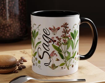 May include: White ceramic mug with a black interior and handle, featuring a botanical illustration of sage plants and the word "Sage" in a script font. The mug sits on a wooden surface with a spoon and a pastry.