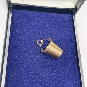 May include: A miniature gold-coloured bucket charm with a handle, set against a blue velvet background within a black box. The bucket has a cylindrical shape and a small handle, suitable for a necklace or bracelet.