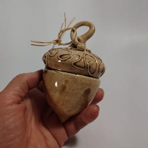May include: A ceramic acorn-shaped container with a glossy tan glaze and speckled finish. The cap features raised, swirling clay detailing and a looped handle at the top, accented with a tied piece of natural raffia string.