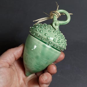 May include: A green ceramic acorn-shaped container with a lid. The lid has a small loop handle and is decorated with small white dots. The container is glazed in a light green color.