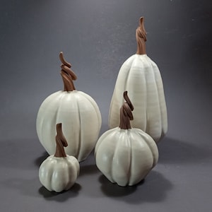 May include: Four white ceramic pumpkins with brown stems. The pumpkins are arranged in a row from largest to smallest.