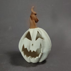 May include: A white ceramic jack-o'-lantern with a carved face featuring triangular eyes and a jagged mouth. The pumpkin has a tall, twisted brown stem extending from the top. The surface is smooth and matte, with deep vertical ridges.