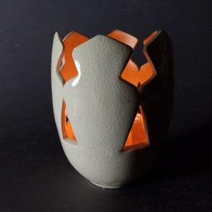 May include: A white ceramic candle holder with a crackled glaze and orange cutouts. The holder is shaped like an egg with a rounded bottom.