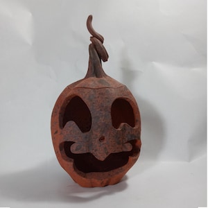 May include: A rustic, orange ceramic jack-o'-lantern with a carved face and a twisted stem. The pumpkin has large eye and mouth cutouts, and a small nose. The surface has a textured, weathered appearance, perfect for Halloween decor.