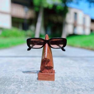 May include: A wooden stand shaped like a face with a pair of black sunglasses resting on the nose.