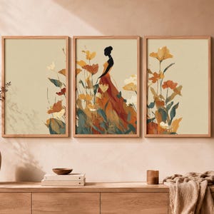 Peut inclure: Un ensemble de trois cadres muraux représentant la silhouette d'une personne en robe fluide au milieu d'illustrations florales orange, jaunes et vertes sur un fond beige clair, présentés dans des cadres en bois clair.