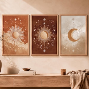 Op de afbeelding: Drie ingelijste hemelse kunstprints. De prints tonen zon- en maanontwerpen in warme tinten oranje, bruin en beige. De zon- en maanillustraties zijn gedetailleerd met sterren en hemelse elementen. De frames zijn gemaakt van lichtgekleurd hout.
