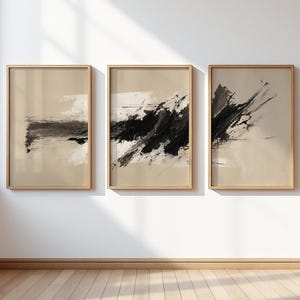 May include: Three vertical rectangular wall art prints in light wood frames. Each print features a beige background with abstract black and white paint strokes, displaying varying textures and dynamic, expressive brushwork across the set.
