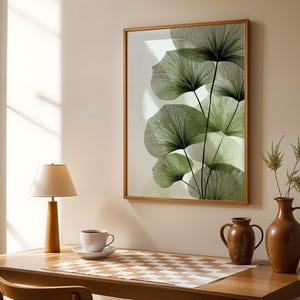 May include: A framed botanical print featuring stylized green leaves with black stems. The artwork is displayed on a beige wall above a wooden desk with a lamp, coffee cup, and decorative vases. The room is bathed in natural light.