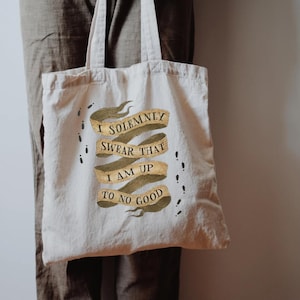May include: Beige canvas tote bag with the text "I SOLEMNLY SWEAR THAT I AM UP TO NO GOOD" in a banner design. The bag has black footprints and a brown handle.