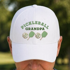 May include: White baseball cap with the text "PICKLEBALL GRANDPA" in green, along with images of pickleball paddles and balls. The cap is a classic style, perfect for outdoor activities.